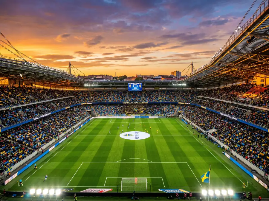 Panoramavy över en fotbollsarena vid solnedgång med spelplanen upplyst och publikhavet runtom